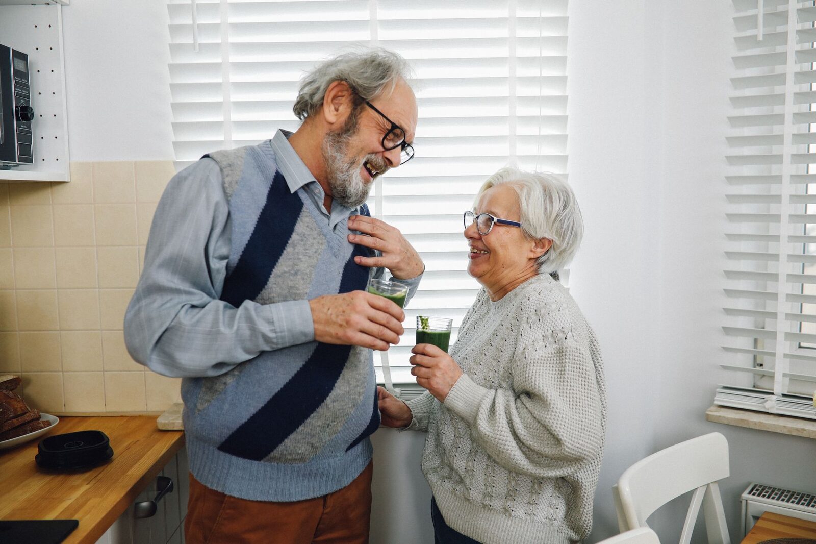 Elderly couple smiling and holding drinks in a cozy kitchen setting.